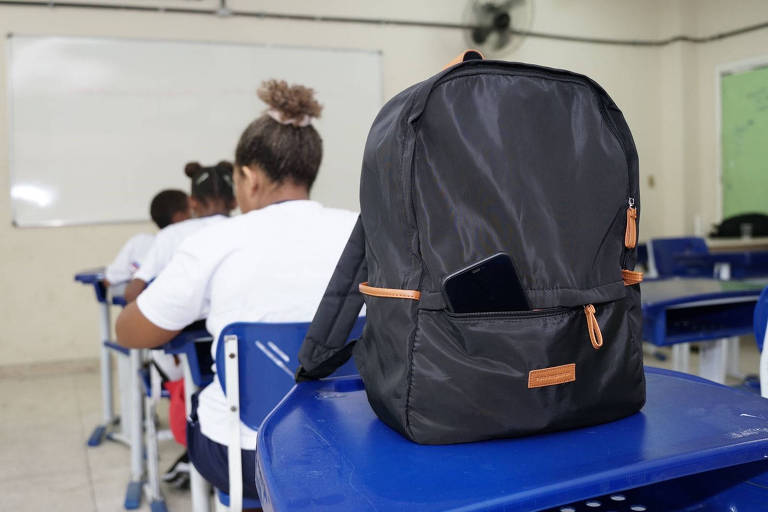 A imagem mostra uma mochila preta com detalhes em laranja, colocada sobre uma mesa em uma sala de aula. Um celular está visivelmente colocado na parte frontal da mochila. Ao fundo, há alunos sentados, de costas, usando camisetas brancas.  A sala possui paredes claras e um quadro branco ao fundo.