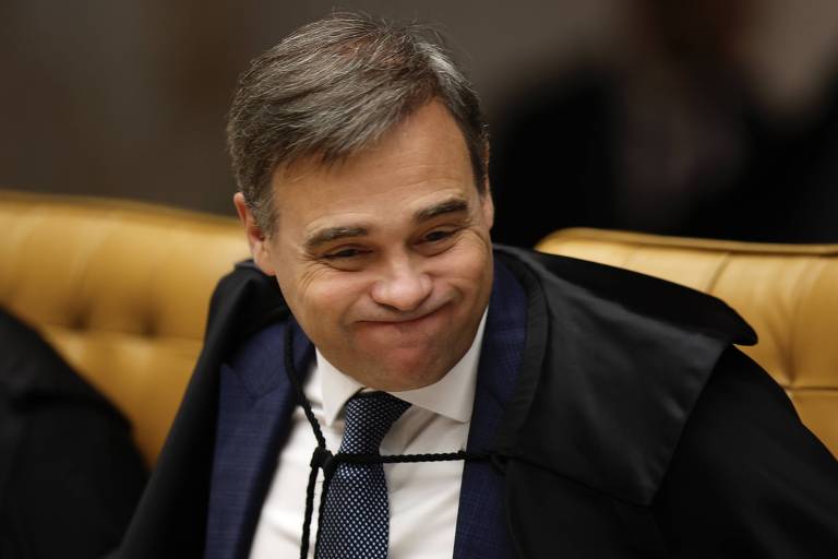 Um homem com cabelo castanho e liso, usando um terno escuro e uma gravata, está sentado em uma cadeira de tribunal. Ele sorri levemente, aparentando estar em um momento descontraído durante uma sessão. O fundo é composto por um painel de madeira e uma cadeira estofada em tom dourado.