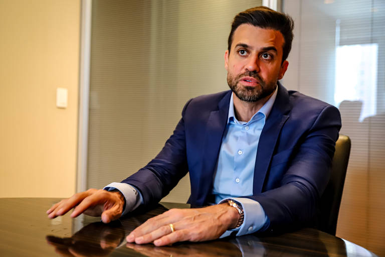 Pablo Marçal, um homem vestindo um blazer azul marinho, senta-se à mesa em uma sala de reuniões, olhando diretamente para a câmera com uma expressão séria e confiante. Suas mãos estão sobre a mesa.