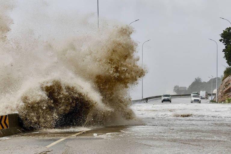 Grandes ondas de cor marrom se chocam contra estrada. Alguns carros podem ser vistos à distância e o céu está nublado