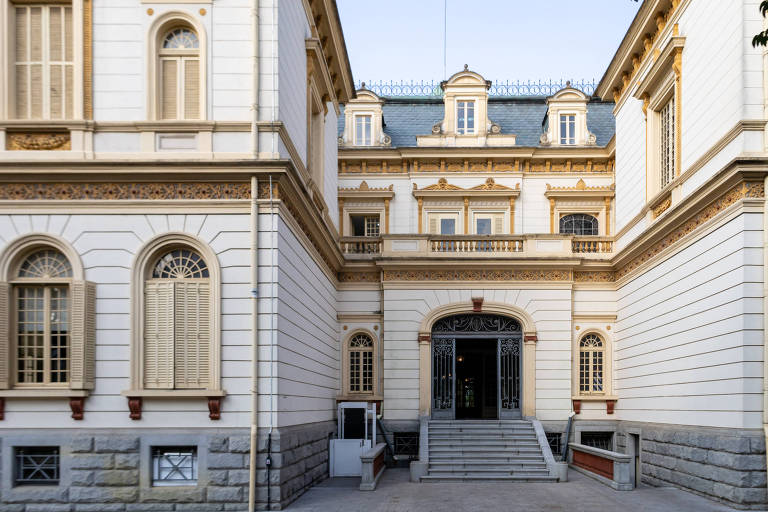 A imagem mostra a fachada de um edifício histórico, com paredes bege claro e detalhes em dourado. A entrada principal é composta por escadas que levam a um grande portal central, flanqueado por janelas com persianas. O telhado é inclinado e possui janelas no sótão. O céu está claro, indicando um dia ensolarado.