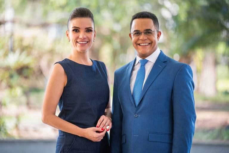 A imagem mostra duas pessoas sorrindo em um ambiente externo. A mulher à esquerda usa um vestido azul escuro e tem cabelo preso. O homem à direita está vestido com um terno azul claro, camisa branca e gravata azul com padrão. Ambos estão em pé, com as mãos visíveis, e o fundo apresenta vegetação desfocada