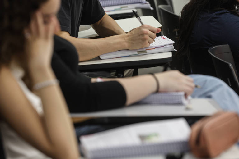 A imagem mostra um grupo de estudantes em uma sala de aula. Em primeiro plano, há duas mãos: uma segurando um caderno e a outra apoiada na mesa. Ao fundo, é possível ver mais estudantes, com alguns cadernos e canetas sobre as mesas. O ambiente parece ser de aprendizado, com foco nas atividades escolares.