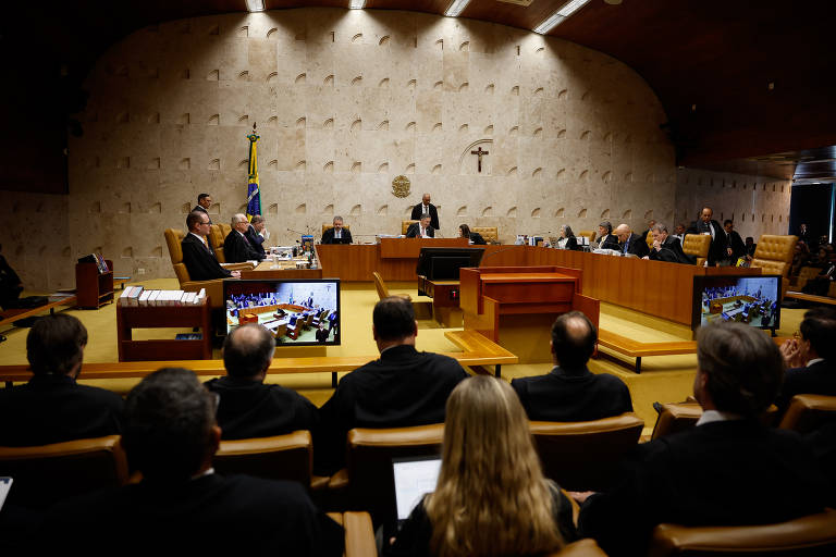 A imagem mostra uma sessão do Supremo Tribunal Federal do Brasil, com vários ministros sentados em uma mesa em formato de 'U'. No fundo, há uma bandeira do Brasil e um crucifixo. Alguns ministros estão em pé, enquanto outros estão sentados, observando. Em frente a eles, há uma mesa com equipamentos de áudio e duas telas exibindo informações. O ambiente é iluminado, com paredes de textura clara.
