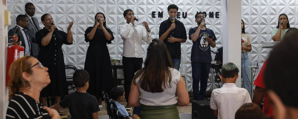 A imagem mostra um grupo de pessoas em uma apresentação musical em um ambiente interno. No fundo, há um palco com várias pessoas cantando e tocando instrumentos. Algumas pessoas estão de pé, enquanto outras estão sentadas em cadeiras. O ambiente é decorado com uma parede texturizada e há um cartaz com a frase 'EM TEU NOME'.