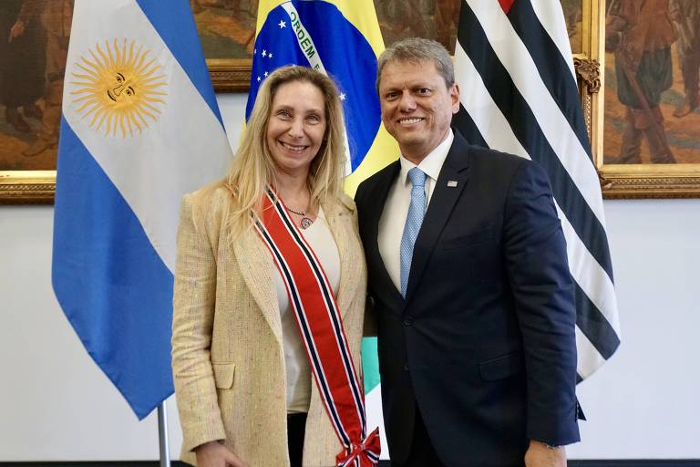 A imagem mostra duas pessoas posando para uma foto em um ambiente interno. À esquerda, há uma mulher com cabelo longo e loiro, vestindo um blazer claro e uma blusa branca, com uma faixa vermelha e branca sobre o ombro. À direita, um homem com cabelo curto e grisalho, usando um terno escuro e uma gravata azul. Ao fundo, estão as bandeiras da Argentina, Brasil e São Paulo, além de uma pintura histórica visível na parede.