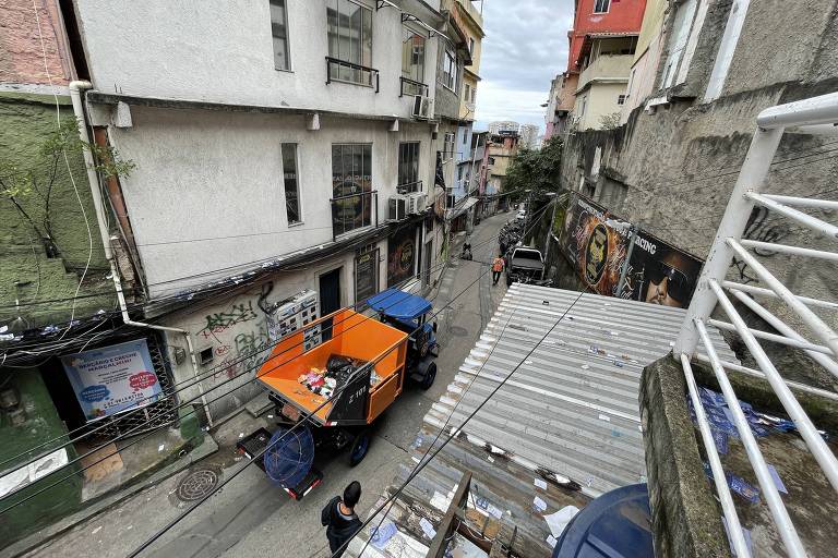 A imagem mostra uma rua estreita em uma área urbana, com prédios de diferentes alturas e cores. Um caminhão laranja está estacionado na rua, e há uma estrutura de telhado de metal visível em primeiro plano. O céu está nublado, e a cena parece ser de um dia comum na cidade.