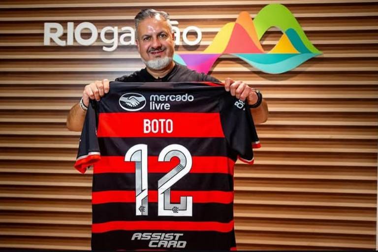 Um homem sorridente está segurando uma camisa de futebol do Flamengo, que é predominantemente preta e vermelha, com o nome 'BOTO' e o número '12' na parte de trás. Ao fundo, há uma parede com o texto 'RIOgaleão' e um design gráfico colorido. O homem usa uma camiseta preta e tem cabelo grisalho e barba.