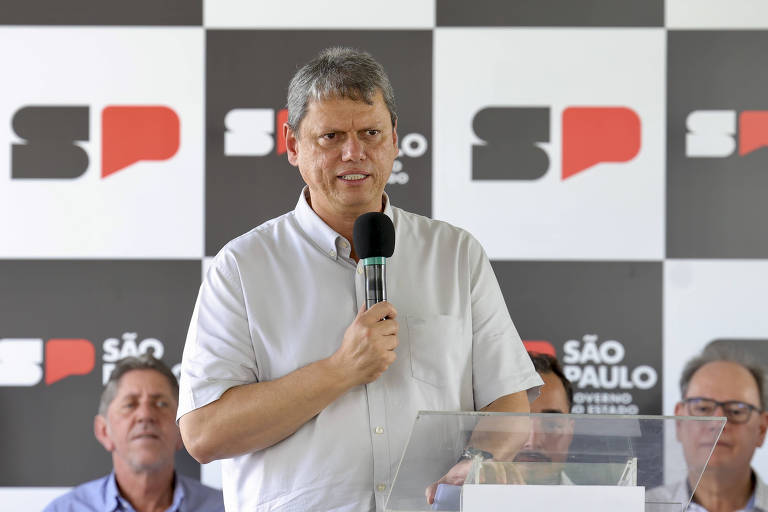 Tarcísio de Freitas, homem de camisa branca, está falando ao microfone em um evento. Ele parece sério e está em frente a um púlpito de vidro. Ao fundo, há um painel com as letras 'SP' em preto e vermelho, e o nome 'SÃO PAULO' em destaque. Dois homens estão sentados atrás dele, um deles olhando para frente e o outro com uma expressão pensativa