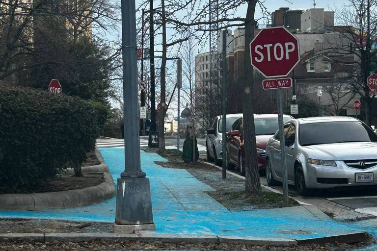 A imagem mostra uma calçada pintada de azul, que é sal, com um sinal de 'STOP' visível ao fundo. Há postes de luz e árvores ao lado da calçada, e alguns veículos estacionados na rua. O céu está nublado.