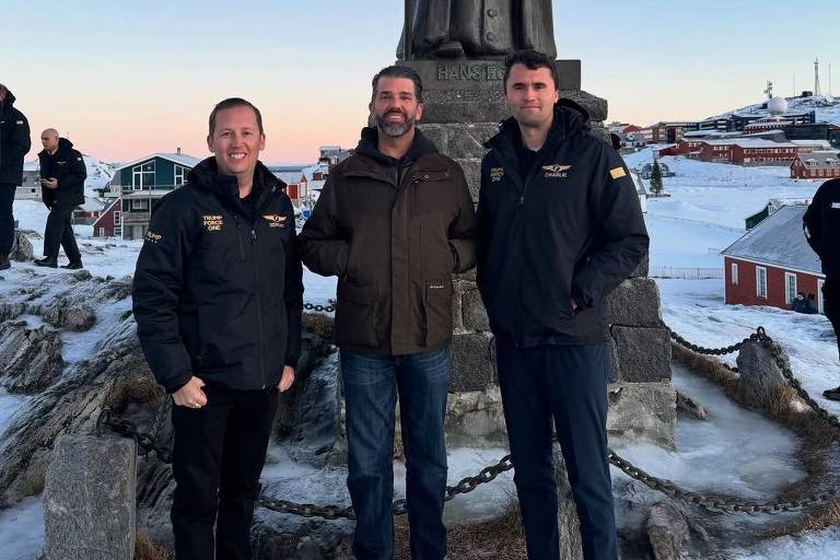 Três homens estão posando para a foto em frente a um monumento em uma área coberta de neve. O céu está claro, com tons de azul e laranja, indicando o início da manhã ou do final da tarde. Os homens estão vestidos com roupas de inverno, e há outras pessoas ao fundo, além de casas e uma paisagem montanhosa