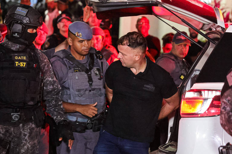 A imagem mostra um homem sendo escoltado por policiais em um ambiente noturno, com luzes vermelhas ao fundo. O homem, que está com as mãos algemadas, usa uma camiseta preta e parece estar sendo levado para um veículo. Dois policiais estão próximos a ele, um deles com um uniforme tático e máscara, enquanto o outro usa um boné e um uniforme da polícia. Há várias outras pessoas e policiais visíveis ao fundo.