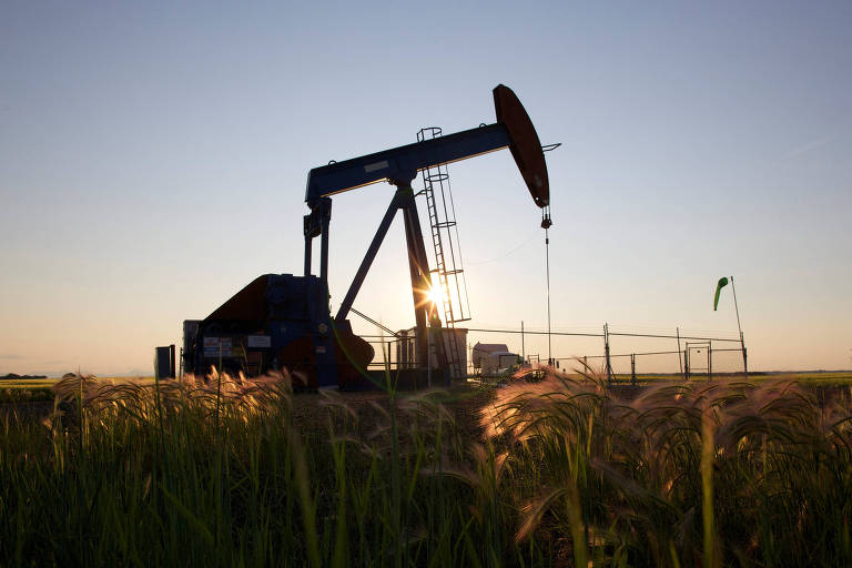 A imagem mostra uma bomba de petróleo em um campo, com o sol se pondo ao fundo. A estrutura da bomba é visível, e há uma cerca ao redor da área de extração. Na frente, há grama alta que se destaca contra o céu claro.