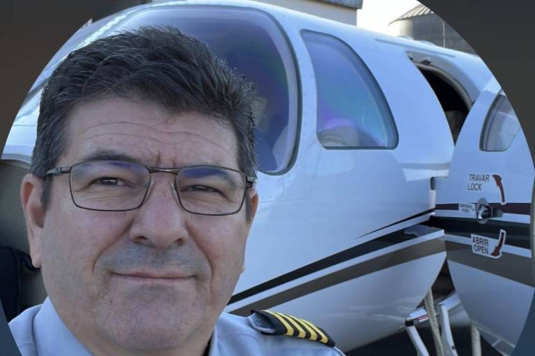 Um homem com cabelo escuro e grisalho, usando óculos e uma camisa de piloto cinza com insígnias amarelas nos ombros, está posando para a foto ao lado de uma aeronave. A parte da aeronave visível é branca com detalhes em preto e há uma porta aberta. O fundo é desfocado, mas parece ser um ambiente de aeroporto.