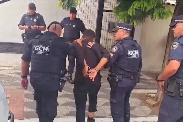 A imagem mostra uma operação policial em que dois policiais estão detendo um homem com as mãos algemadas. O homem está vestido de preto e é acompanhado por outros policiais que observam a situação. Ao fundo, há uma parede e algumas árvores. A cena parece ocorrer em uma área urbana.