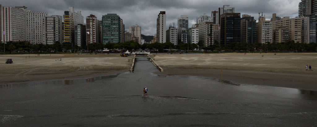 A imagem mostra uma vista panorâmica de uma cidade com prédios altos e modernos ao fundo, sob um céu nublado e escuro. No primeiro plano, há uma área de areia molhada e a água que chega à praia por um canal. Há um casal passeando pela água em frente ao canalA atmosfera é sombria devido às nuvens.