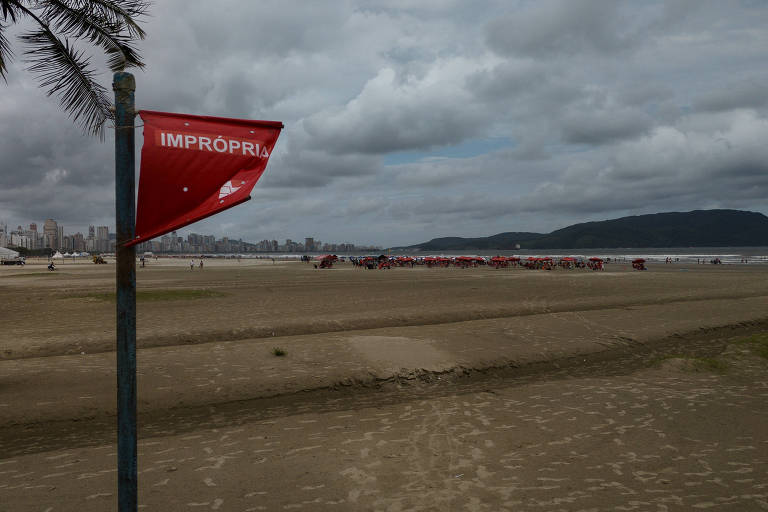 A imagem mostra uma praia com uma bandeira vermelha hasteada, indicando que a água é imprópria para banho. Ao fundo, há uma cidade com prédios e um céu nublado. Na areia, algumas pessoas e guarda-sóis vermelhos são visíveis.