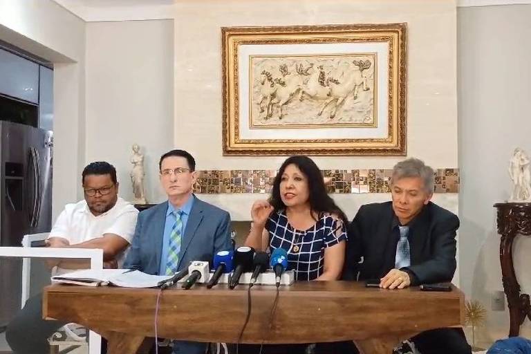 A imagem mostra uma coletiva de imprensa com quatro pessoas sentadas em uma mesa. À esquerda, um homem de camiseta branca. No centro, um homem de terno azul claro e gravata. Ao lado, uma mulher com blusa de bolinhas e cabelo solto, que parece estar falando. À direita, um homem de terno escuro. Ao fundo, uma parede com uma obra de arte e uma mesa decorativa.