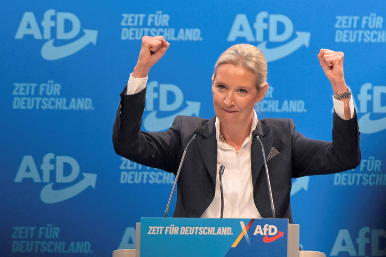 Uma mulher em pé, com os braços levantados em sinal de celebração, fala em um evento político. Ela está vestida com um blazer escuro e uma blusa clara. Ao fundo, há um painel azul com o logotipo de um partido político e uma frase que diz, em alemão, 'A hora da Alemanha'.