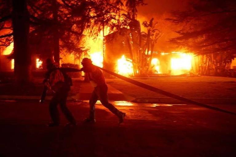 Foto mostra a silhueta de um bombeiro correndo com uma mangueira nos ombros em meio a incêncio em uma casa arborizada. 