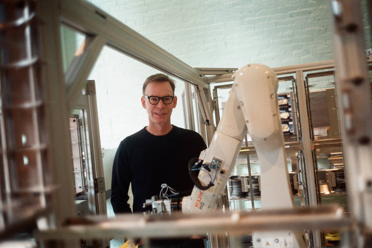 A imagem mostra um homem usando óculos e uma camiseta preta, posicionado em um ambiente industrial com um braço robótico ao seu lado. O robô está em uma estrutura metálica, e o fundo é composto por equipamentos e maquinário.