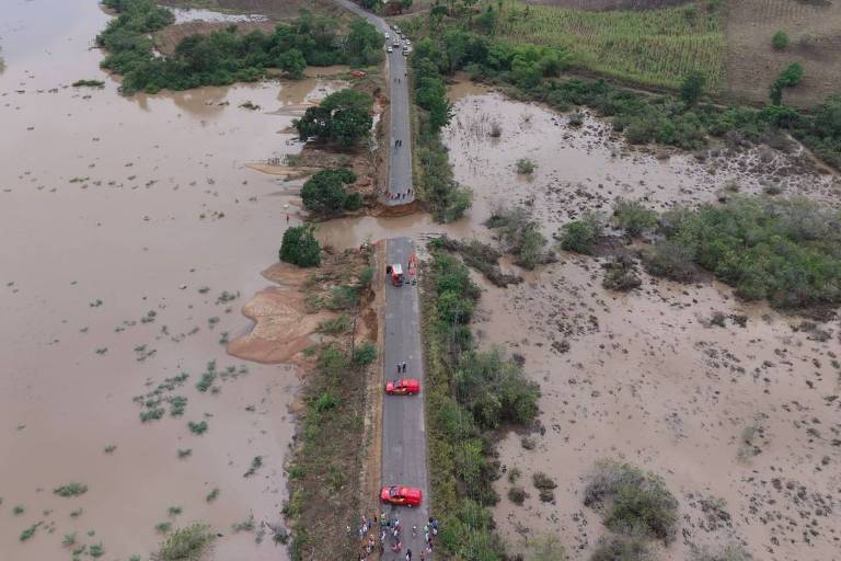 A imagem mostra uma estrada que se estende por uma área alagada, com água em ambos os lados. O cenário inclui campos verdes e uma paisagem montanhosa ao fundo. Há alguns veículos visíveis na estrada, e a água parece ter invadido as margens, criando uma grande extensão de alagamento.