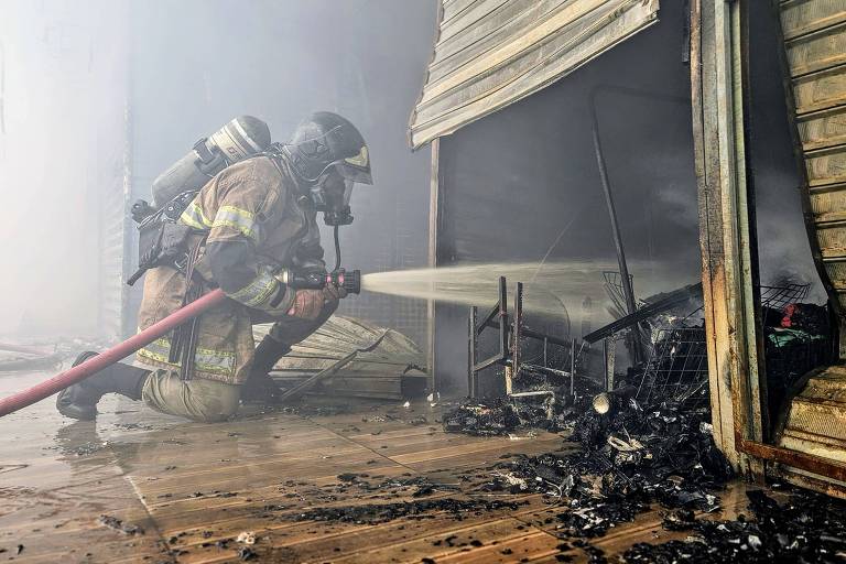 Um bombeiro está agachado, usando um equipamento de proteção, enquanto combate um incêndio em um ambiente com fumaça densa. Ele direciona um jato de água para uma área que parece ter sido queimada, com restos carbonizados visíveis no chão. A estrutura ao fundo é uma parede de madeira com tábuas expostas.