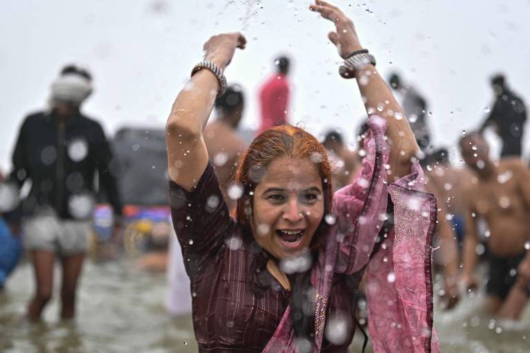 Uma mulher com cabelo castanho claro e uma blusa escura está sorrindo e levantando os braços enquanto água espirra ao seu redor. Ela usa um lenço rosa e está em um ambiente aquático, onde várias outras pessoas estão visíveis ao fundo, algumas parcialmente submersas na água.