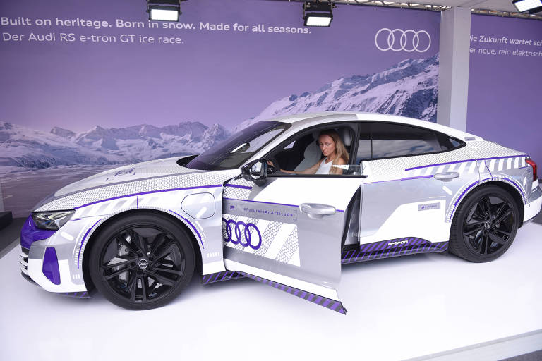 A imagem mostra um carro da marca Audi, modelo elétrico, estacionado em um evento. O veículo é predominantemente branco com detalhes em roxo e possui uma porta aberta. Dentro do carro, há uma pessoa visível. Ao fundo, há uma parede com o logotipo da Audi e texto em português que menciona a marca e o modelo do carro.