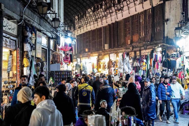 A imagem mostra um mercado noturno em Damasco, com várias barracas e lojas iluminadas. Há muitas pessoas caminhando pelas ruas, algumas paradas para olhar produtos. As lojas vendem roupas, acessórios e outros itens. O ambiente é movimentado e vibrante, com uma arquitetura tradicional visível ao fundo.
