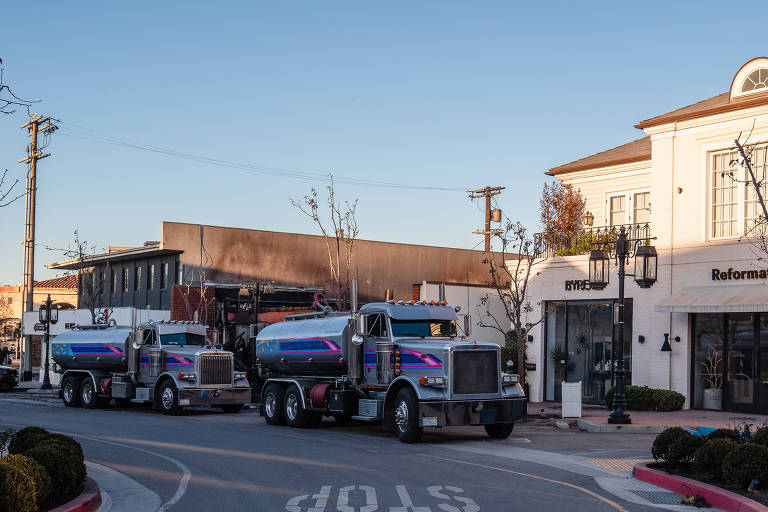 A imagem mostra dois caminhões-tanque estacionados em uma rua comercial. O primeiro caminhão, à esquerda, é prateado com detalhes em azul e rosa. Ao fundo, há um edifício com uma fachada escura, possivelmente danificada, e um outro edifício branco com janelas e uma placa que diz 'Reformation'. A rua é pavimentada e há uma sinalização de 'STOP' visível no chão. Árvores sem folhas estão presentes ao longo da calçada.