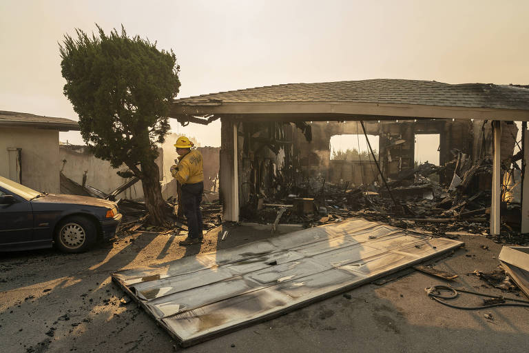 A imagem mostra um bombeiro em uma área devastada por incêndio, ao lado de um carro azul e uma árvore. O fundo revela uma estrutura de casa que foi severamente danificada, com paredes queimadas e escombros visíveis. O céu está nublado, indicando fumaça ou poluição, e há uma porta de garagem caída no chão.