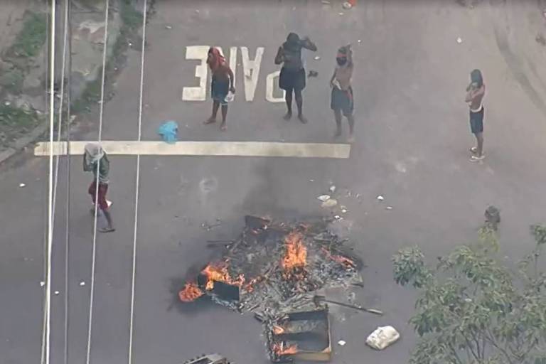 A imagem mostra uma rua com um grupo de pessoas em pé, algumas delas em frente a uma fogueira. Há uma barricada feita de objetos queimados no centro da rua. O ambiente parece tenso, com fumaça visível. Ao fundo, há vegetação e uma linha de postes de eletricidade.