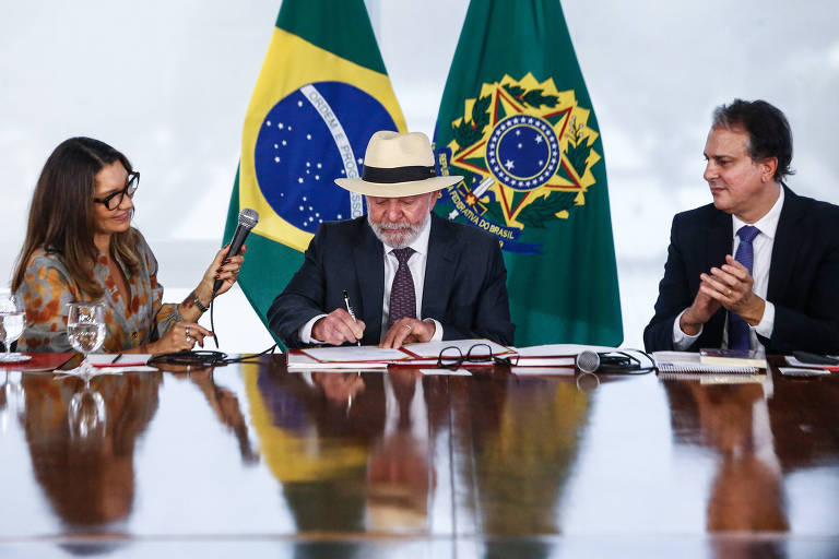 A imagem mostra uma reunião oficial com três pessoas sentadas à mesa. À esquerda, Janja, uma mulher com óculos e cabelo solto segura um celular, observando. No centro, um Lula, com um chapéu branco, assina documentos, enquanto à direita, Camilo Santana aplaude. Ao fundo, estão as bandeiras do Brasil e um símbolo do governo.