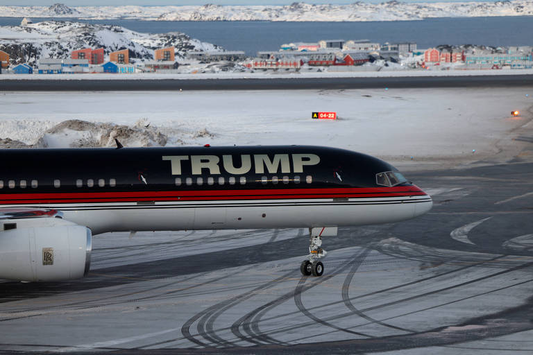 Um avião comercial com a inscrição 'TRUMP' na fuselagem está estacionado em uma pista coberta de gelo. O avião possui uma pintura preta com detalhes em vermelho e uma bandeira dos Estados Unidos na cauda. Ao fundo, há uma paisagem de cidade com edifícios e uma área de água, sob um céu em tons de rosa e azul.