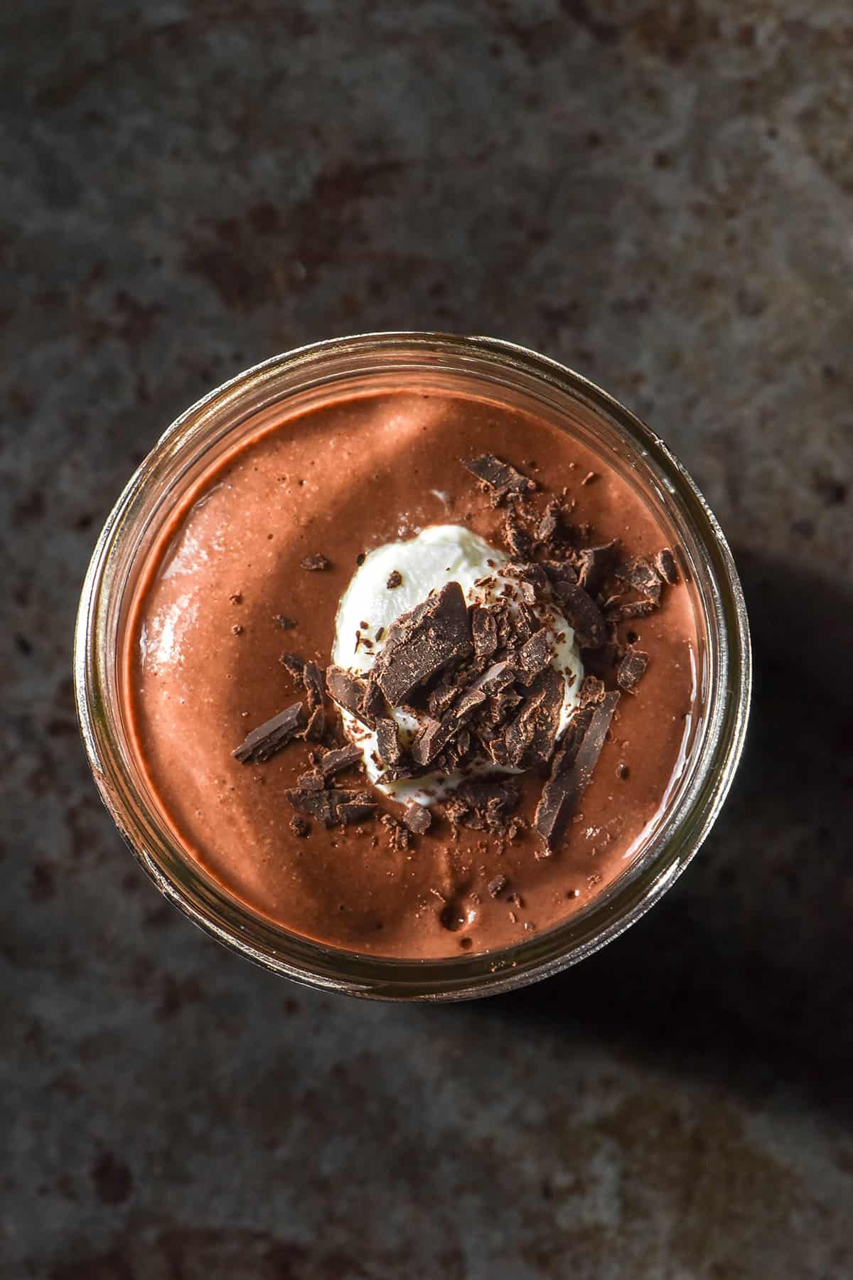 An aerial image of a chocolate protein chia pudding in a glass jar topped with a spoonful of high protein yoghurt and a sprinkling of chocolate shards. The pudding sits atop a dark grey metal backdrop.