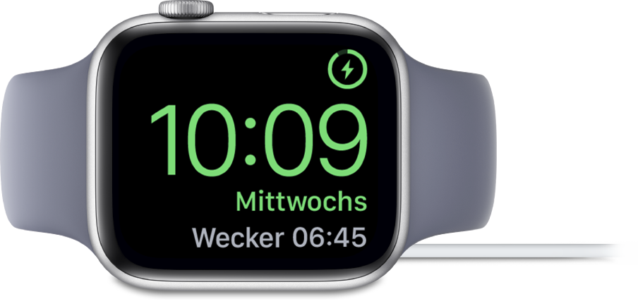 Eine auf die rechte Seite gedrehte Apple Watch, an die das Ladegerät angeschlossen ist und auf deren Display oben rechts das Ladesymbol, darunter die aktuelle Uhrzeit und die Uhrzeit für den nächsten Wecker angezeigt werden.