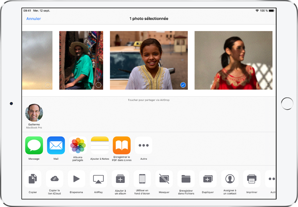 L’écran Partage avec des photos affichées en haut ; l’une d’elles est sélectionnée, comme indiqué par la coche blanche dans un rond bleu. Le rang sous les photos présente les amis avec lesquels vous pouvez les partager à l’aide d’AirDrop. D’autres options de partage sont proposées en dessous, notamment, de gauche à droite : Messages, Mail, Albums partagés, Ajouter à Notes, Enregistrer le PDF dans Livres et Autre. Le rang du bas contient les boutons suivants : Copier, Enregistrer dans Fichiers, Copier le lien iCloud, Diaporama, AirPlay, Ajouter à un album, Utiliser en fond d’écran, Masquer, Dupliquer, Assigner à un contact, Imprimer et Autre.
