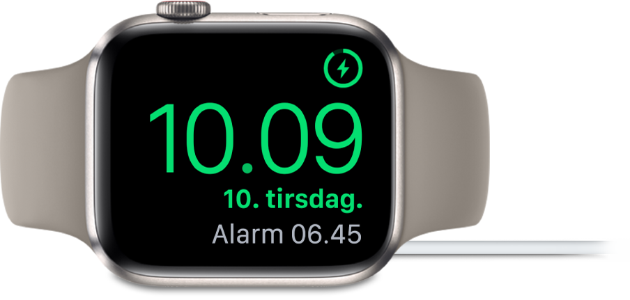 Et Apple Watch, der ligger på siden og er sluttet til opladeren, hvor skærmen viser opladersymbolet øverst til højre, det aktuelle klokkeslæt derunder og klokkeslættet for den næste alarm.