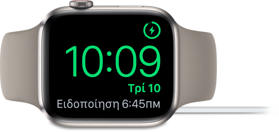 Ένα Apple Watch τοποθετημένο στο πλάι του και συνδεδεμένο στον φορτιστή του. Στην οθόνη εμφανίζεται το σύμβολο φόρτισης στην πάνω δεξιά γωνία, η τρέχουσα ώρα από κάτω η ώρα του επόμενου ξυπνητηριού.