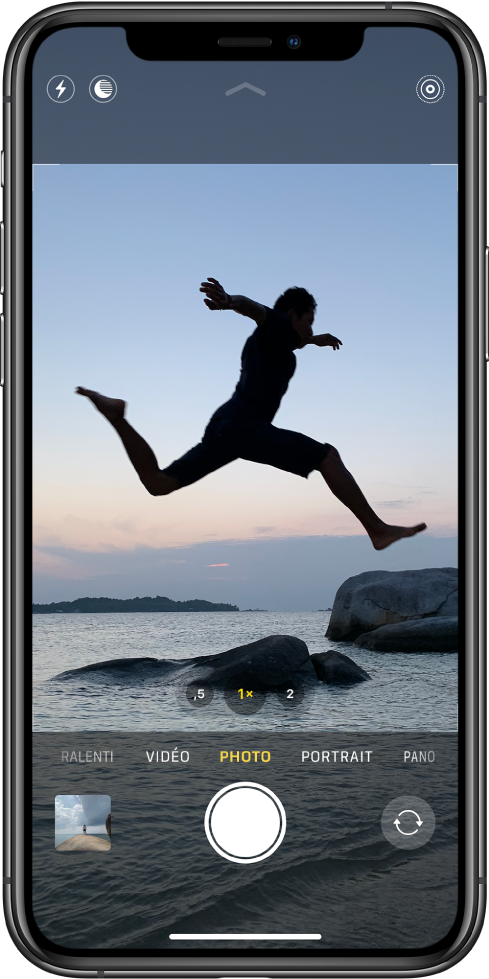 L’écran Appareil photo en mode Photo, avec les autres modes à gauche et à droite sous le visualiseur. Les boutons pour le flash, le mode nuit et le mode Live Photo se situent en haut de l’écran. En dessous des modes de l’appareil photo figurent, de gauche à droite, une vignette permettant d’accéder aux photos et vidéos, le bouton Obturateur et le bouton « Changer de caméra ».