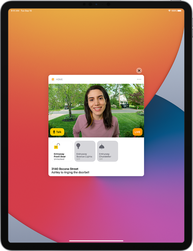 Na zaslonu iPada je obvestilo aplikacije Home. Prikazuje sliko osebe pred vhodnimi vrati z gumbom Talk na levi. Spodaj so gumbi pripomočkov za vhodna vrata in luči v hodniku. Pod gumbi pripomočkov se prikaže domači naslov skupaj z besedilom »Ashley is ringing the doorbell«. Zgoraj desno od obvestila je gumb Close.