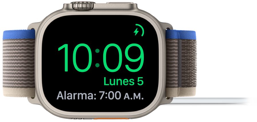 Un Apple Watch colocado de lado y conectado al cargador, con la pantalla mostrando el símbolo de carga en la esquina superior derecha, la hora actual debajo y la hora de la siguiente alarma.