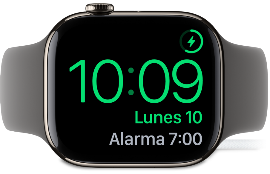 Un Apple Watch colocado de lado y conectado al cargador, en cuya pantalla se muestra el símbolo de carga en la esquina superior derecha, la hora actual debajo y la hora de la siguiente alarma.