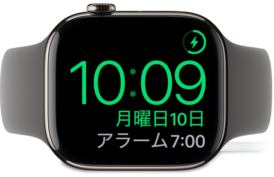 横向きに置かれ、充電器に接続されているApple Watch。画面には、右上隅に充電中のマーク、その下に現在時刻と、次のアラーム時刻が表示されています。