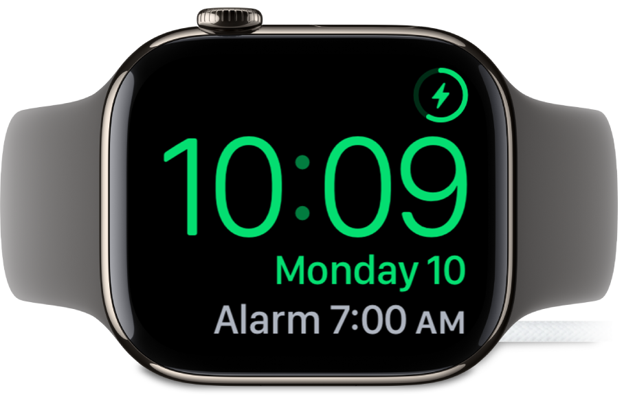 Apple Watch ustawiony na boku i podłączony do ładowarki. W prawym górnym rogu ekranu widoczny jest symbol ładowania, niżej wyświetlana jest bieżąca godzina, a dalej znajduje się czas najbliższego alarmu.