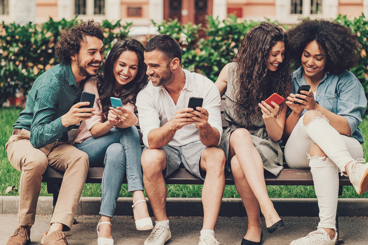 Happy young adults sitting on a park bench looking at their phones. 