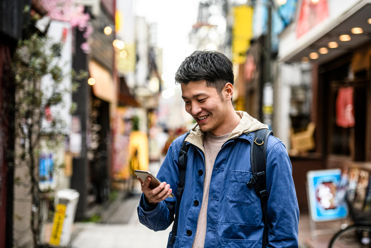 Young man in a blue jacket reading a text on his cell surrounded by street vendors. 