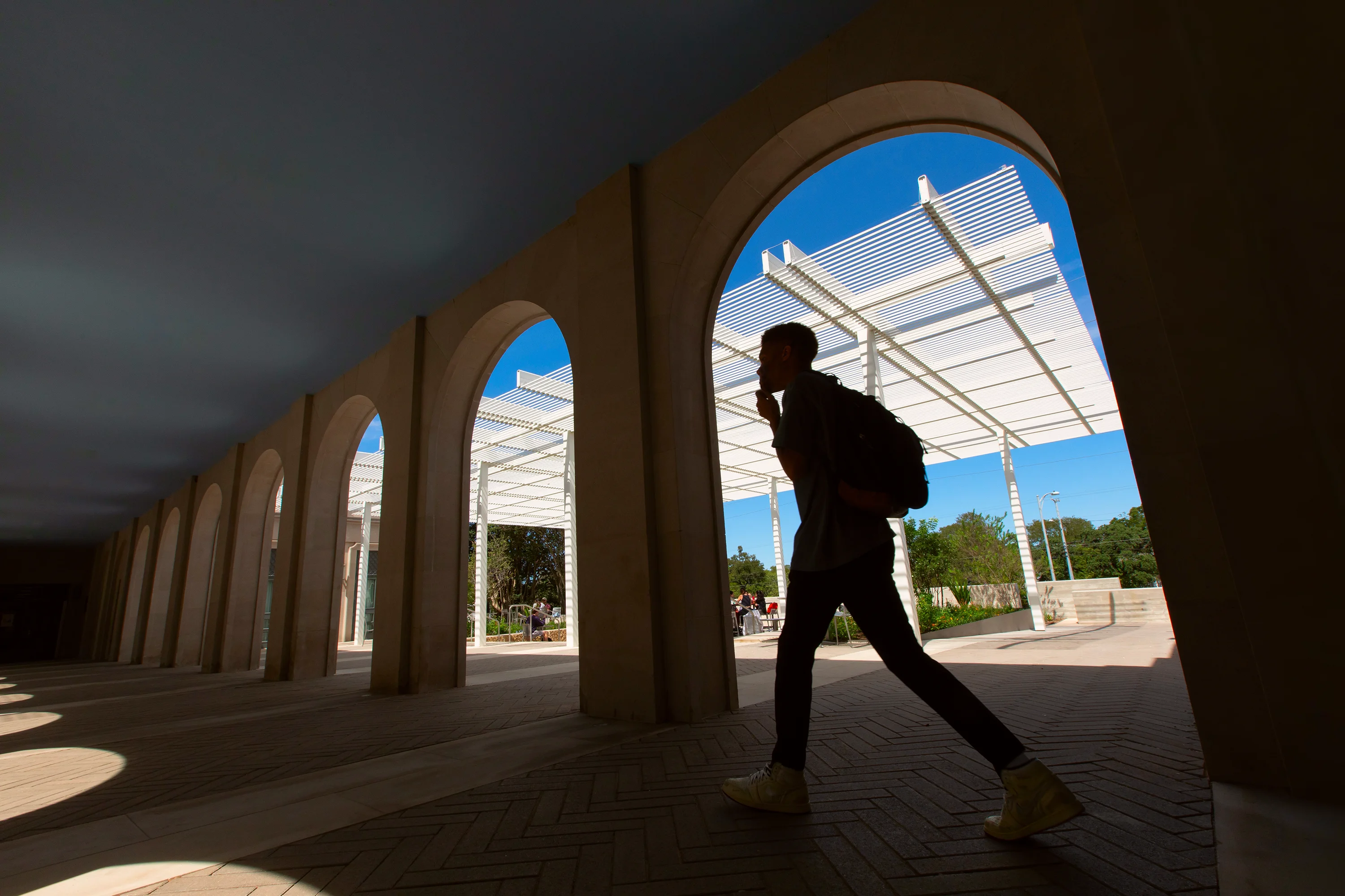 A student walks across a shaded arched breezeway, heavily silhouetted by a beautiful blue sky