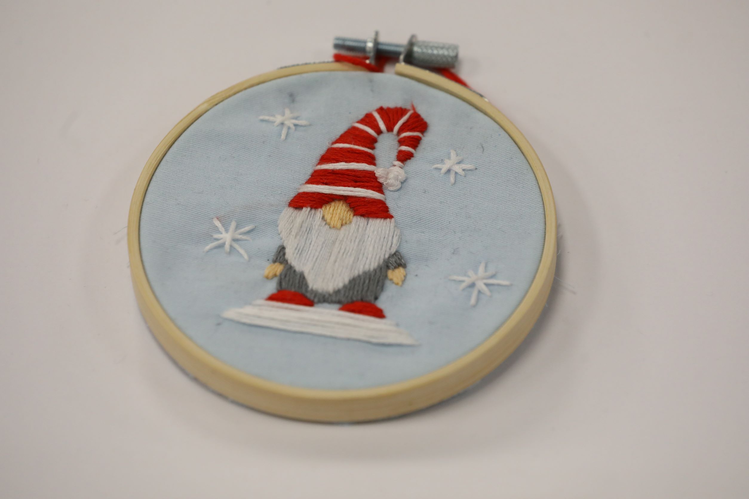 Hand Embroidered Gnome wearing a red and white hat with a white beard standing on ice with snowflakes around on a hoop